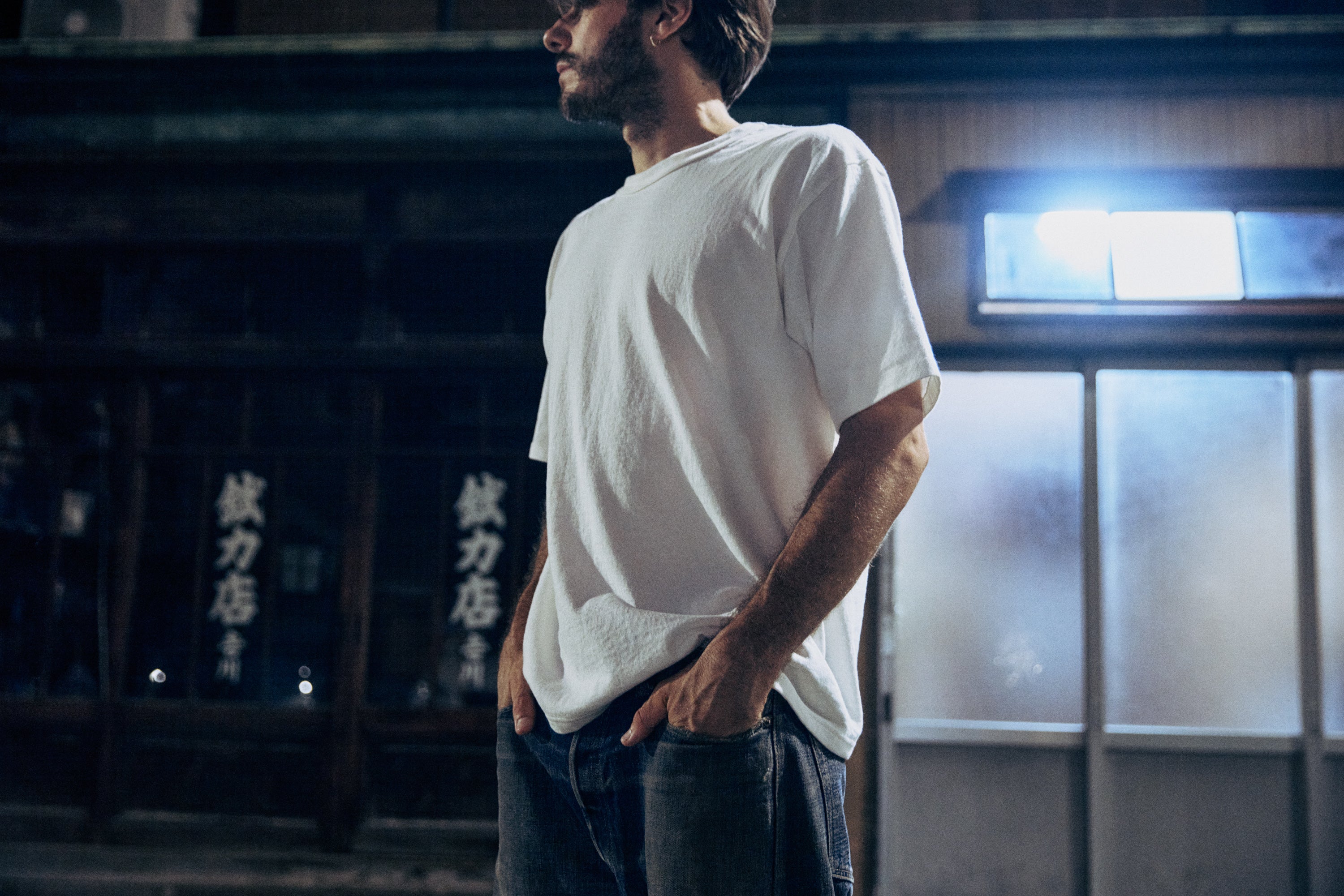 Man wearing a white t-shirt and dark pants standing in a dimly lit room with Japanese text on the wall.
